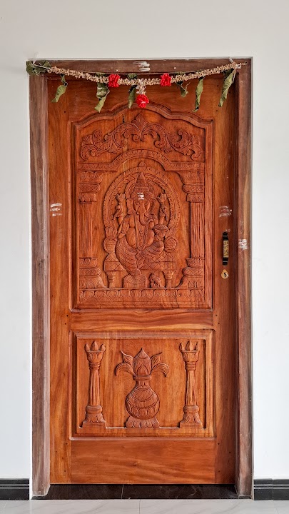 Carved timber entrance door featuring Lord Ganesha on a throne and sacred kalash with ceremonial lamps